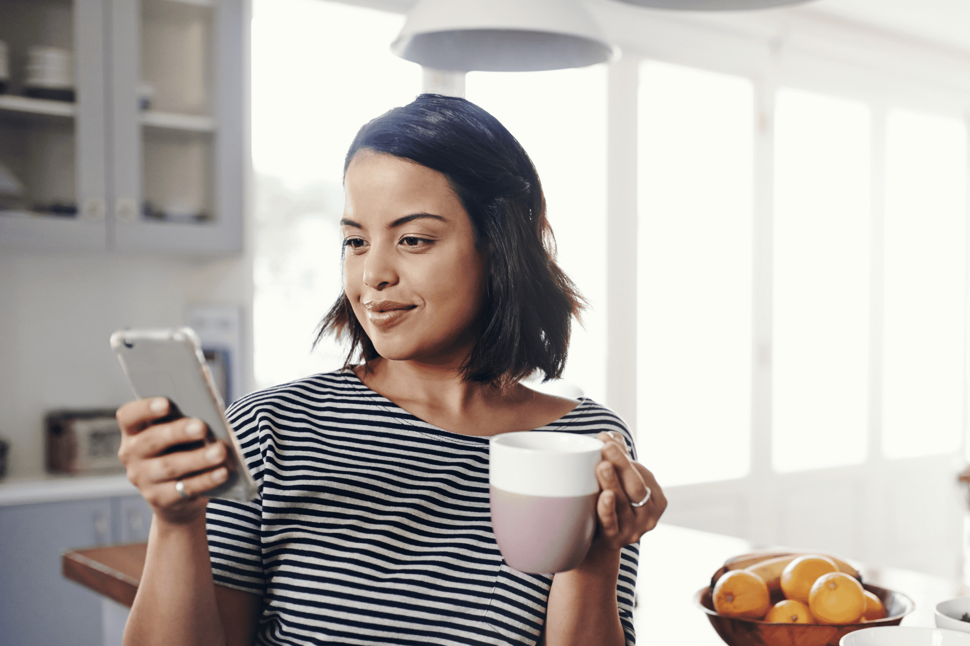 Woman holding phone and coffee in kitchen.