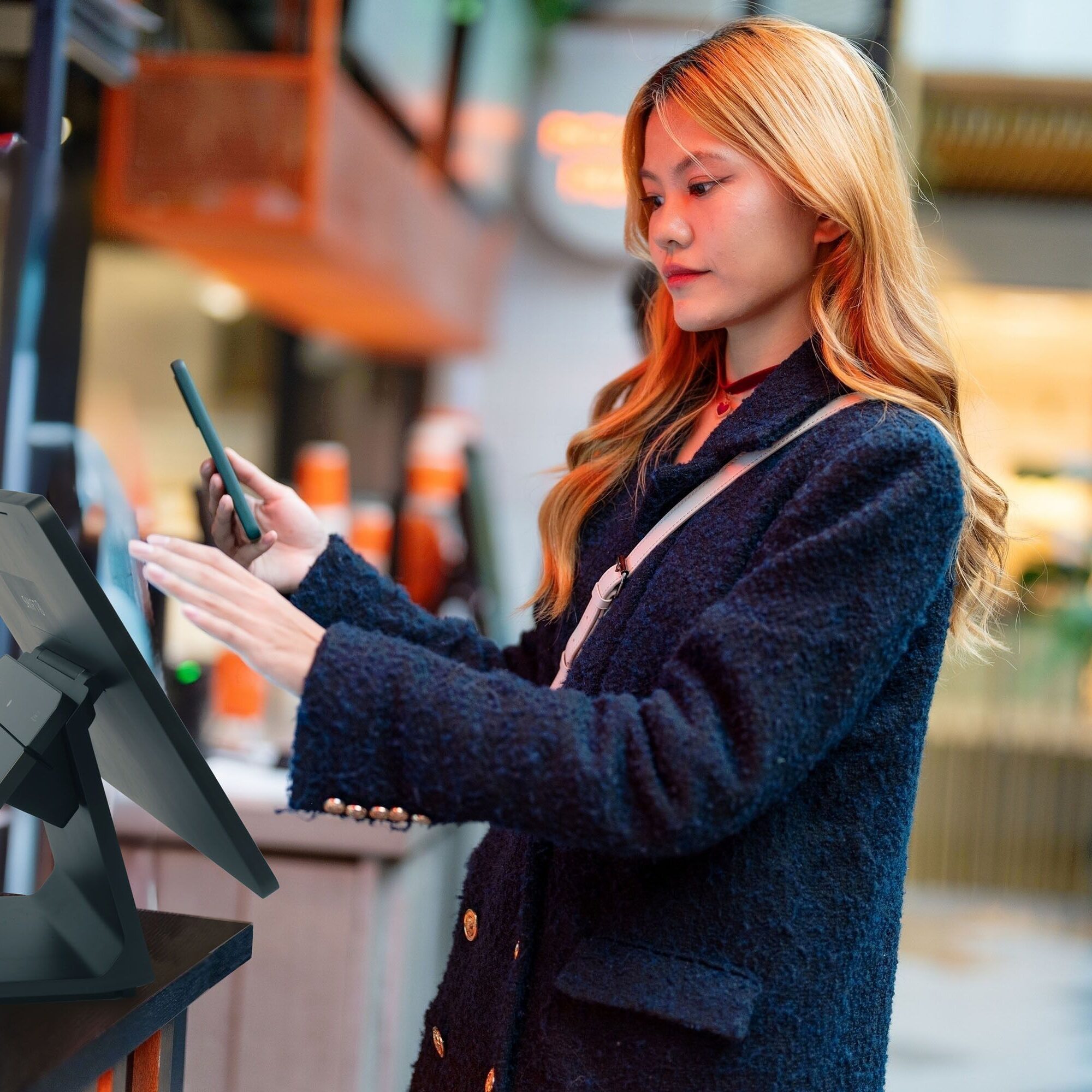 Woman using phone at a touchscreen kiosk.
